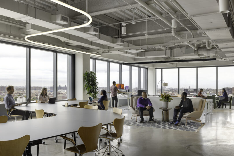 Modern office space with employees working at desks and in lounge areas, featuring large windows and an urban view