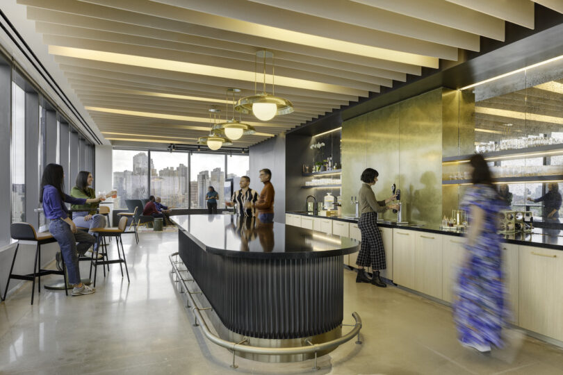 Modern office break room with people socializing and getting coffee, featuring stylish bar seating, pendant lighting, and panoramic city views