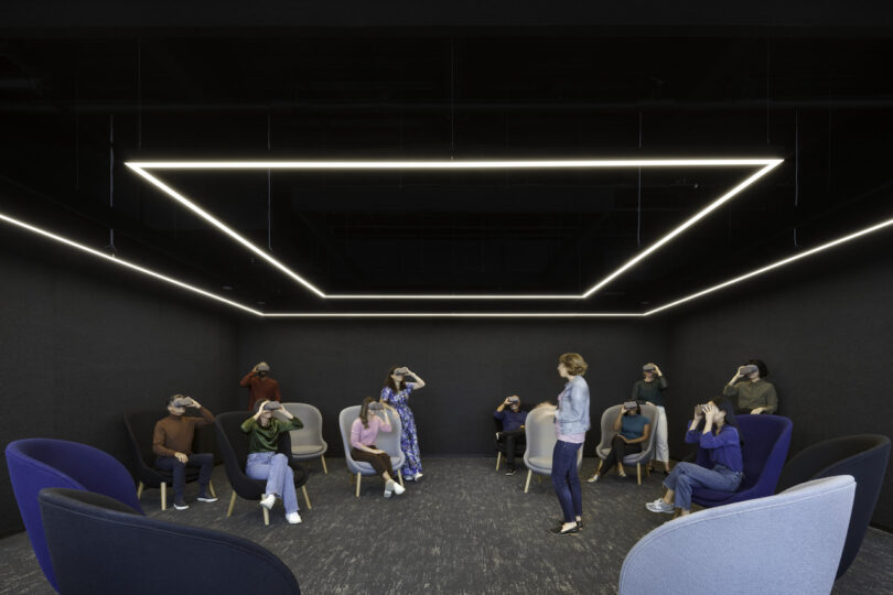 A modern office break room with triangular ceiling lights, where a diverse group of employees wearing vr headsets is guided by a woman standing