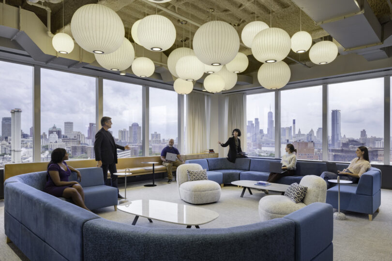 Modern office lounge with people sitting and standing, featuring large windows with city skyline views and spherical pendant lights