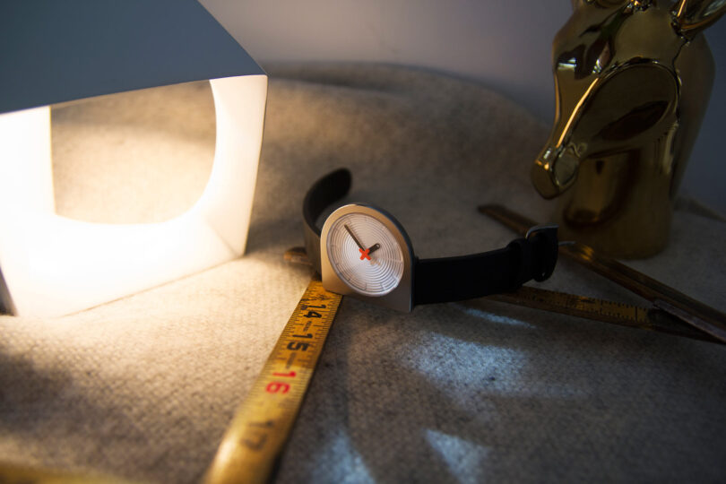 D-shaped analog wristwatch with black band set across fabric upholstery near modern lamp, abstract metallic animal head sculpture, and measuring stick