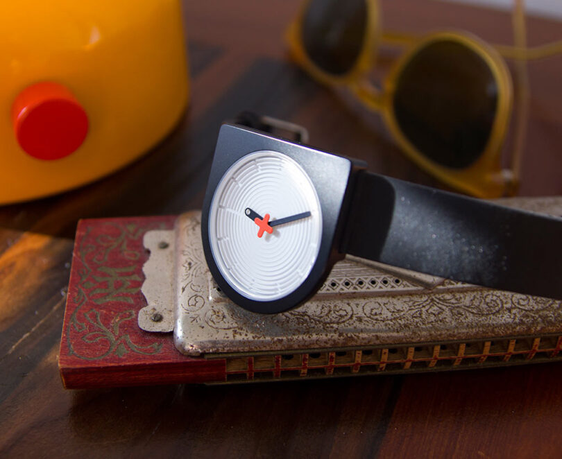 A modern D-shaped wristwatch with a black strap, resting on an ornate metallic box beside sunglasses and a yellow teapot.