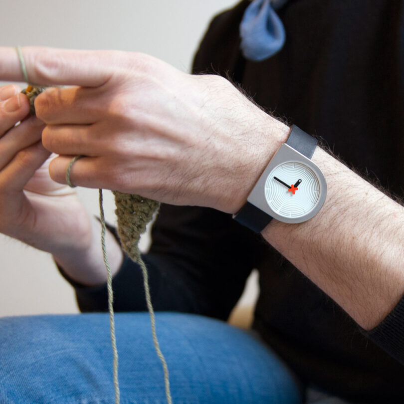 A man wearing a D-shaped wristwatch on his left wrist knits with a green yarn.