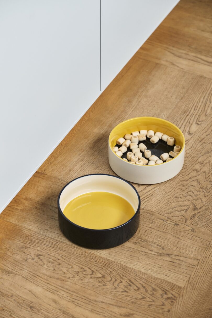two white, yellow, and black dog bowls on the floor with food and water in them