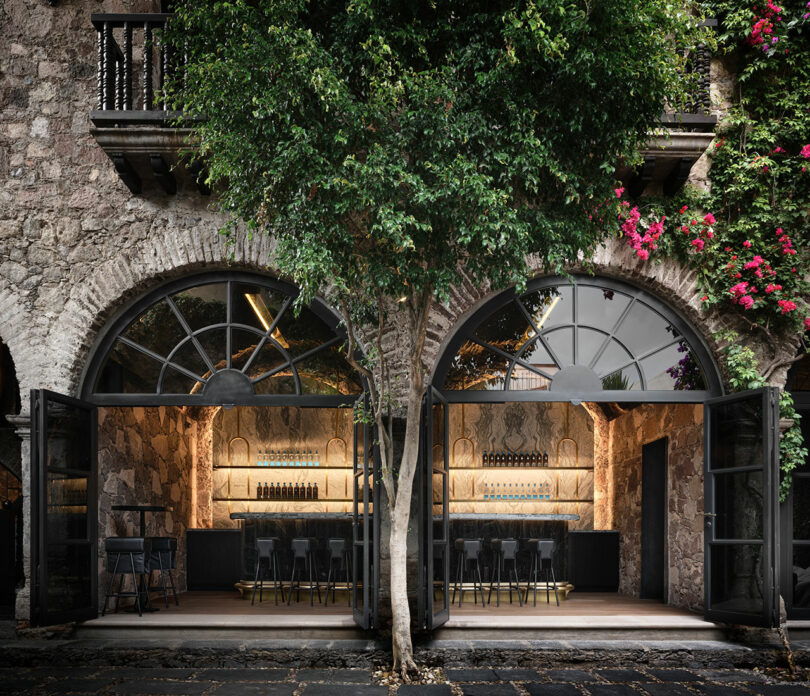 Outdoor view of bar with arched glass doors, stone walls, and a tree, with stools and a lit interior visible.