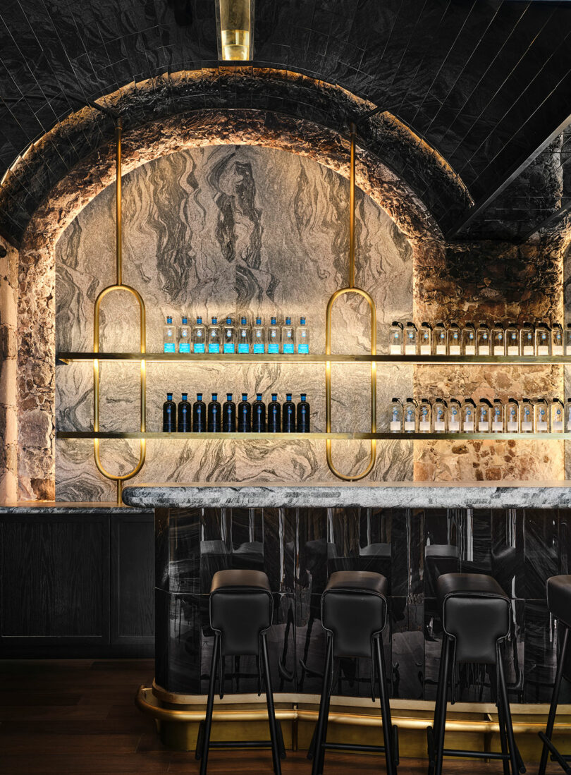 Elegant bar interior with arched stone walls, marble countertop, and shelves of Casa Dragones bottles backlit by golden lighting.