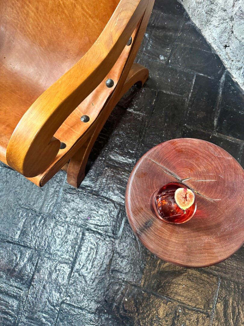 A Casa Dragones cocktail with a lime slice on a round wooden table next to a wooden chair with metal bolts, on a textured black floor.