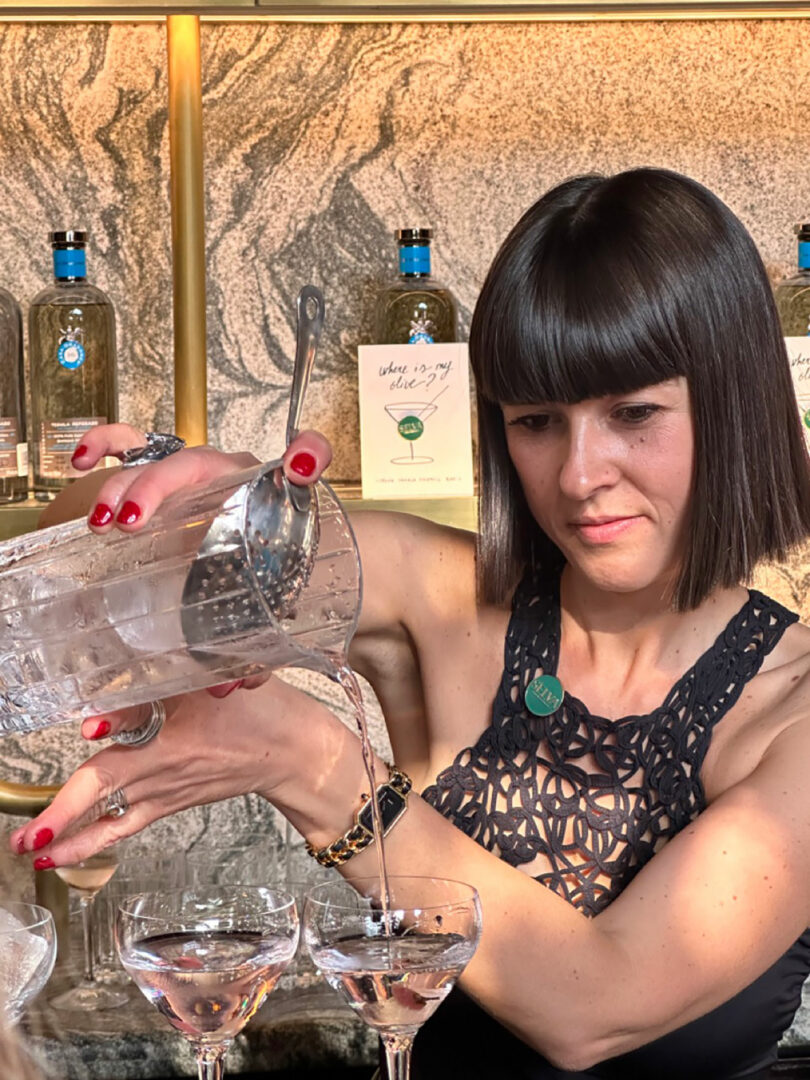 A woman meticulously pours a clear beverage into cocktail glasses at a bar, surrounded by bottles. She has short dark hair and wears a black sleeveless top.
