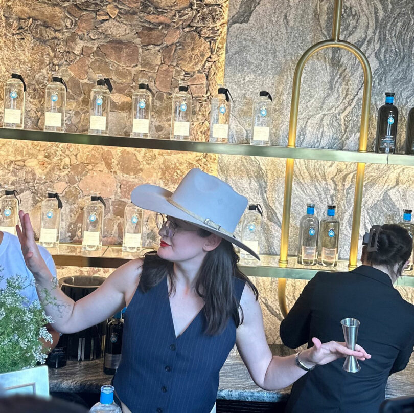Woman in a white hat and dark vest gesturing while talking at a bar with shelves of Casa Dragones bottles and a stone wall backdrop.