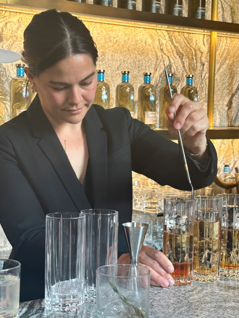 A bartender in a black blazer preparing Casa Dragones drinks with a bar spoon, standing behind a counter lined with bottles and glasses.