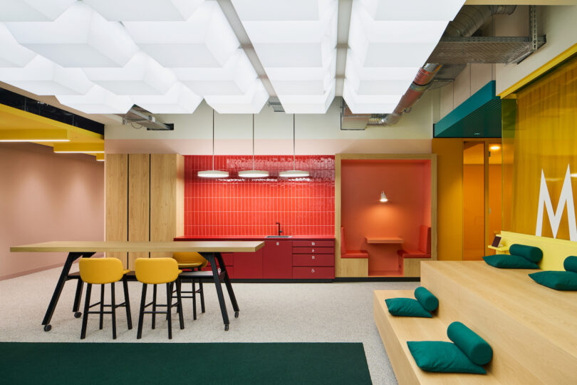 Modern office break area with colorful interior design, featuring a communal table, kitchenette, and seating nook.