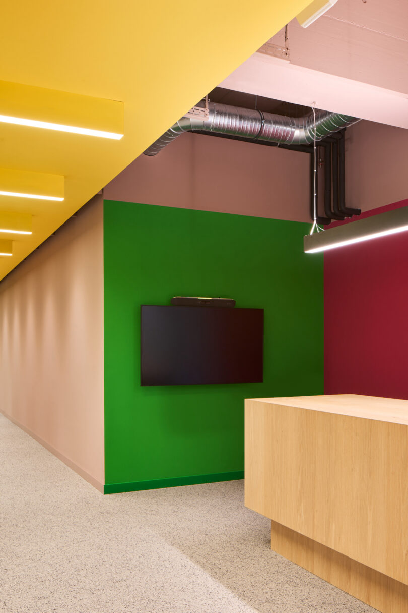 A modern office space featuring a green wall with a mounted flat-screen tv, intersected by a bold yellow beam, accompanied by a wooden desk and pink accent lighting.