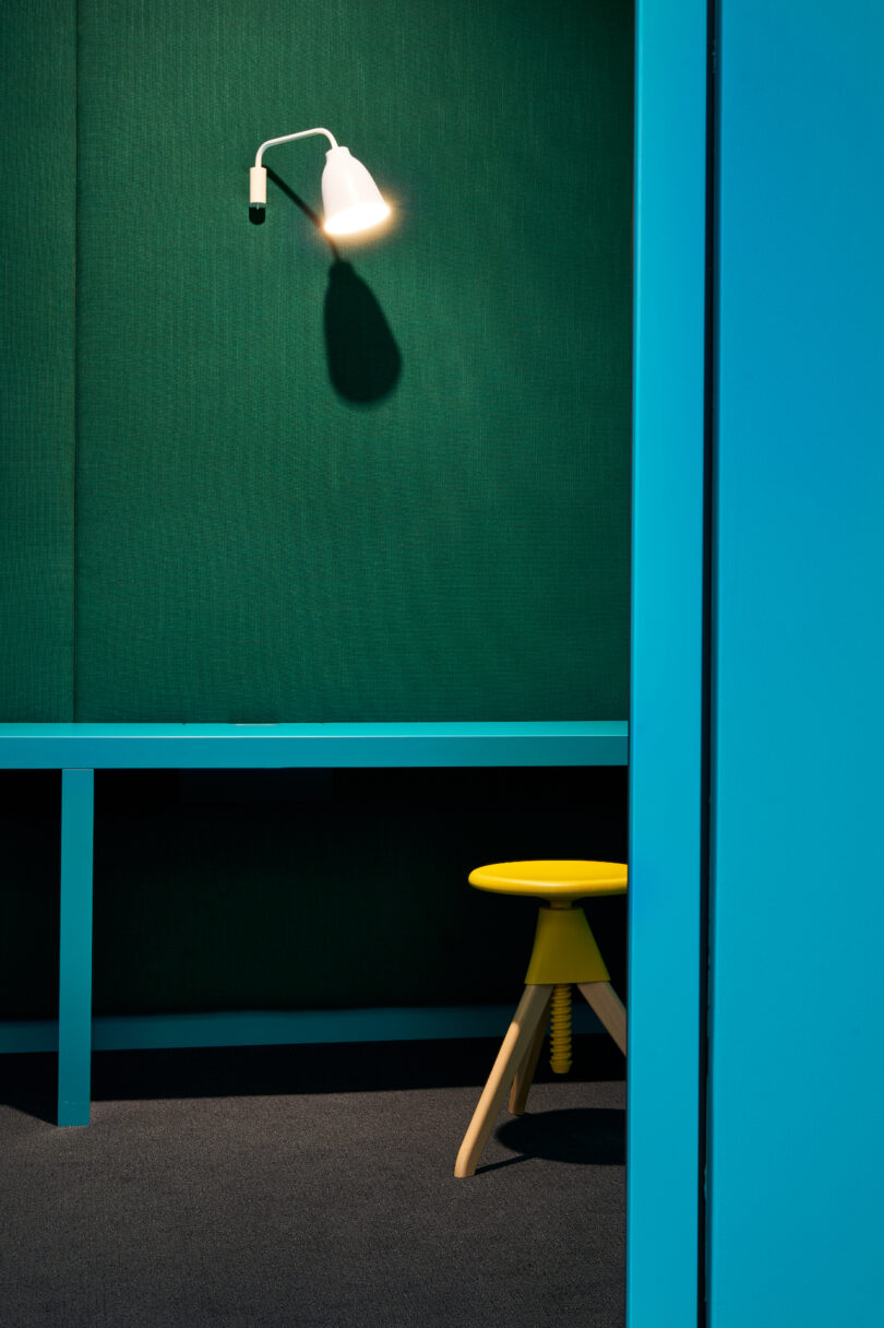 A single wall lamp casts a warm glow on a textured green wall beside a blue-framed nook with a yellow stool.