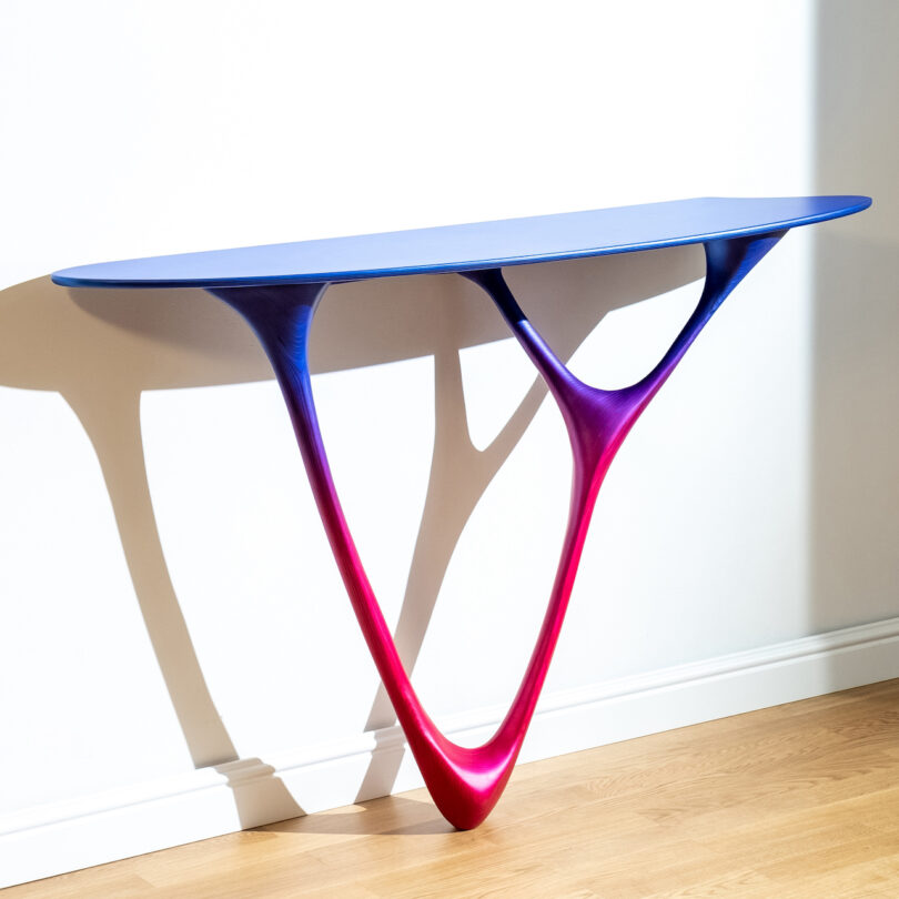 A modern table with a gradient color design transitioning from blue to red, against a white wall on a wooden floor.