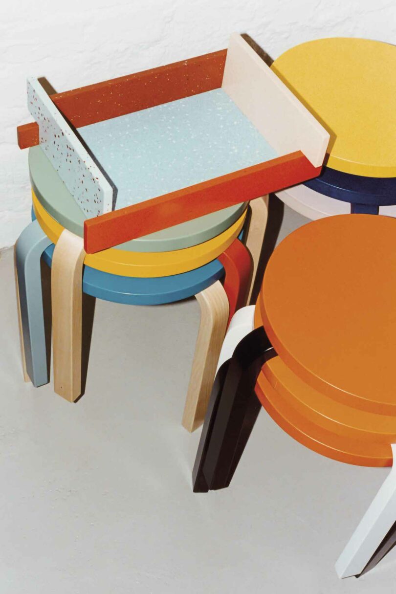 A stack of colorful, modern chairs with varied designs and shapes, neatly arranged in a corner against a white wall.
