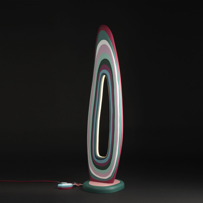 A modern sculptural lamp with layered multicolored rings stands illuminated against a dark background, featuring a visible power cord.