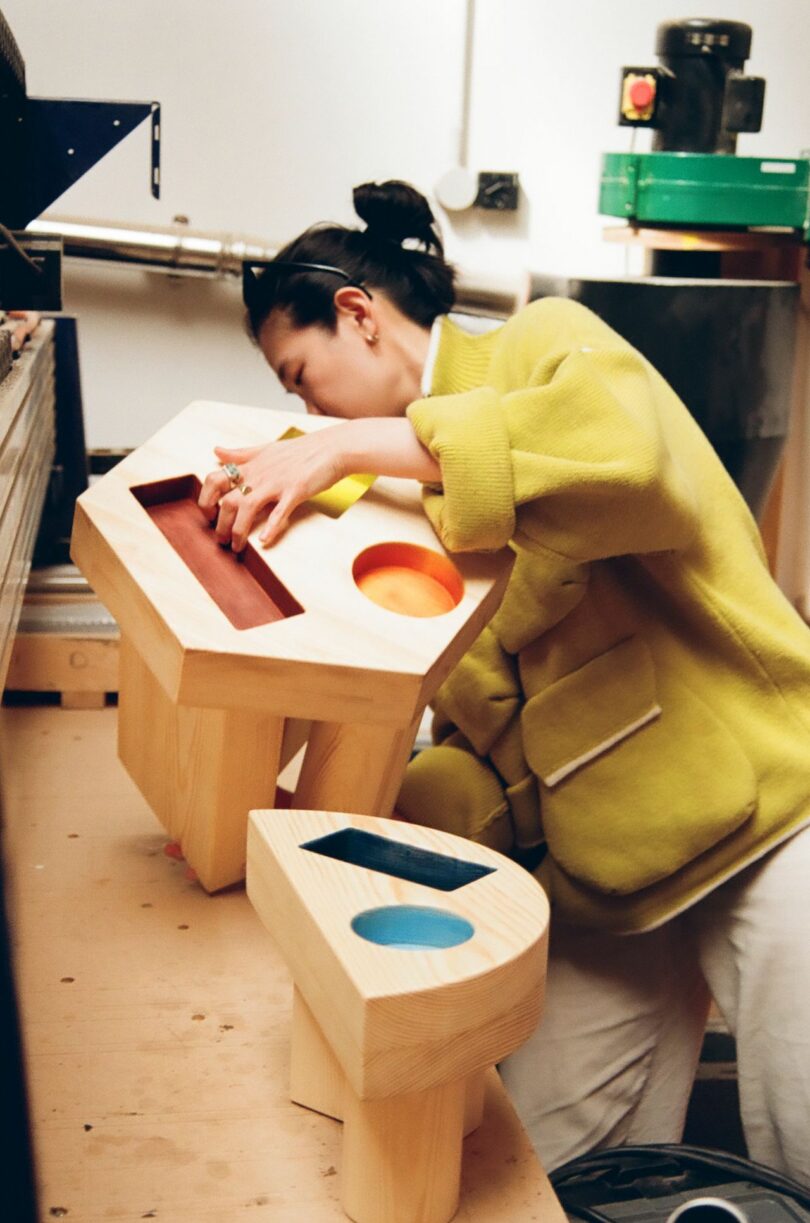woman holding a wood side table with colorful geometric cut outs on a work bench