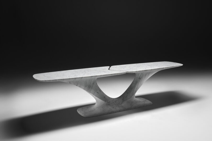 Modern marble table with a unique v-shaped base and a split, elongated oval top, highlighted against a black background.