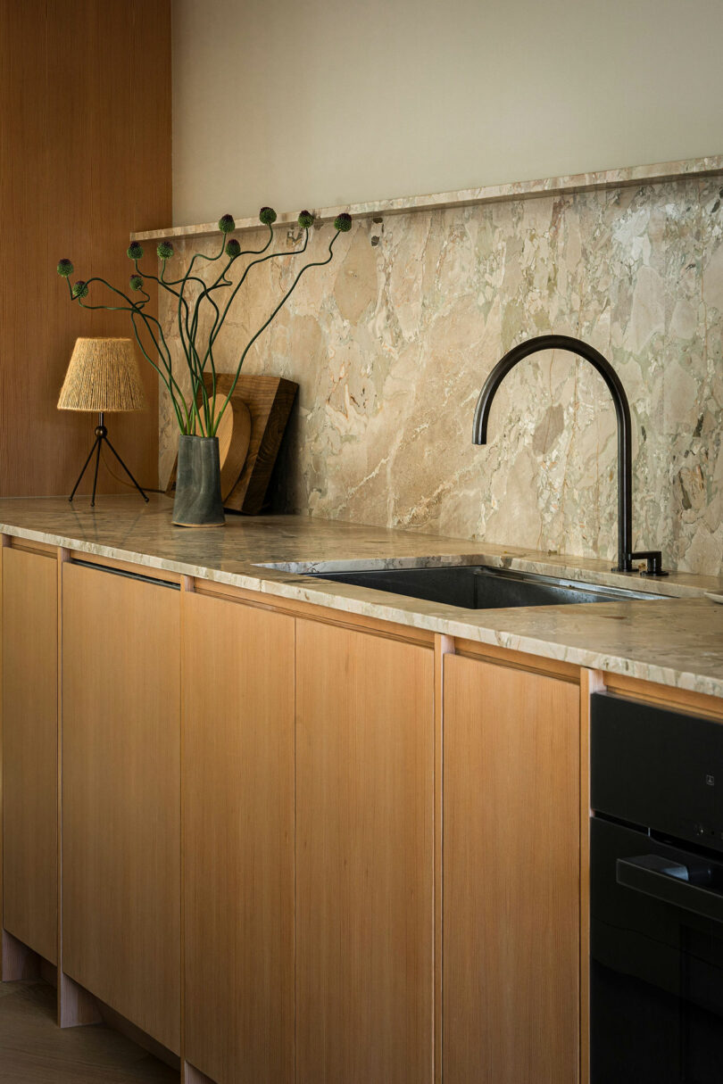A modern kitchen with a beige marble countertop and backsplash, wooden cabinets, a black faucet, and a small lamp with a potted plant on the counter.