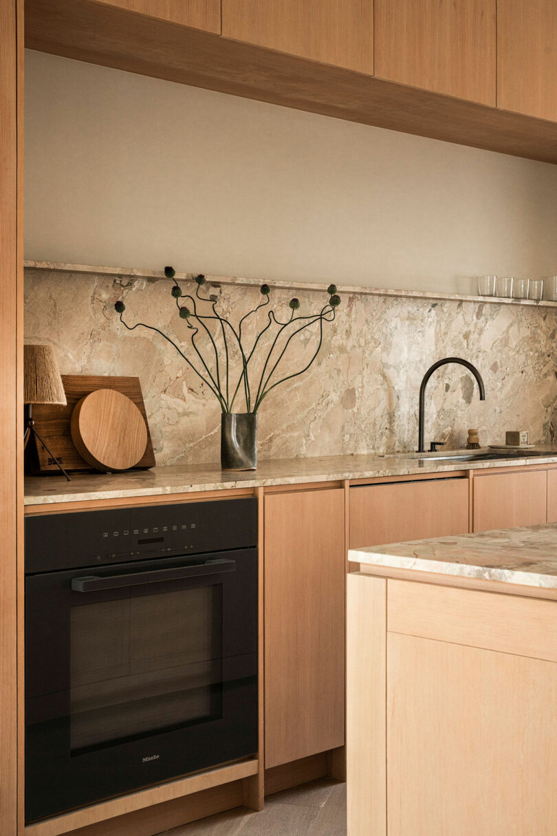 A modern kitchen with light wood cabinets, a marble countertop and backsplash, a black oven, a black faucet, and minimal decor including a vase with branches and a wooden cutting board.