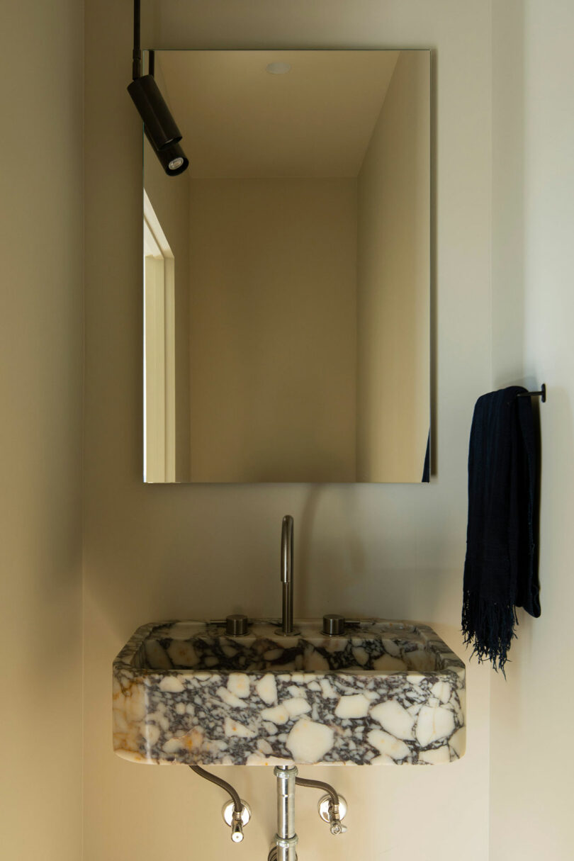 A bathroom sink with a marble countertop, a sleek faucet, a wall-mounted mirror, a black hand towel on a towel ring, and a black ceiling light fixture.