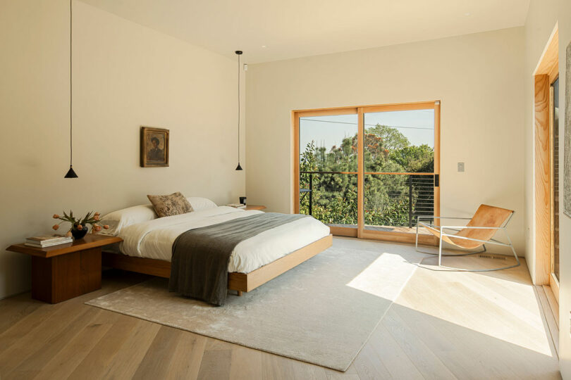 A minimalist bedroom featuring a low wooden bed with white bedding and a gray throw, flanked by two nightstands. A large window with a green view and a modern chair complete the room.