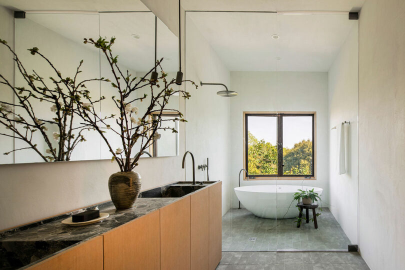 Modern bathroom with a freestanding bathtub, a large mirror, a black marble countertop, wooden cabinetry, and a vase with branches. A window provides natural light and a view of greenery.