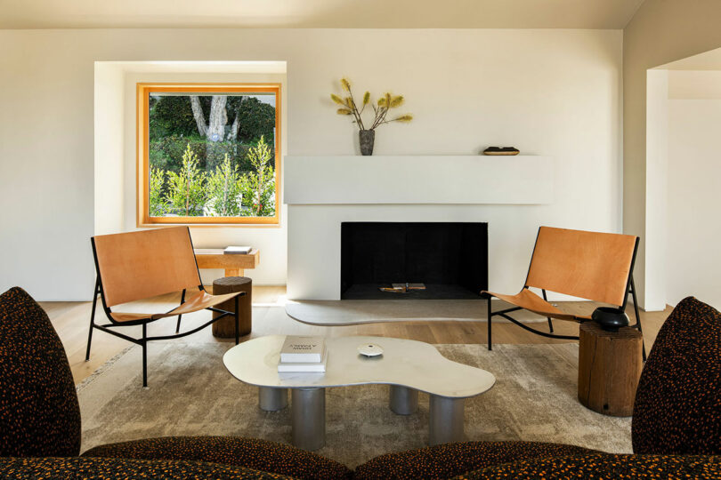 A minimalist living room with two tan leather chairs, a white fireplace, a sculptural coffee table, a window with a scenic view, and sparse decor including a vase and small objects.