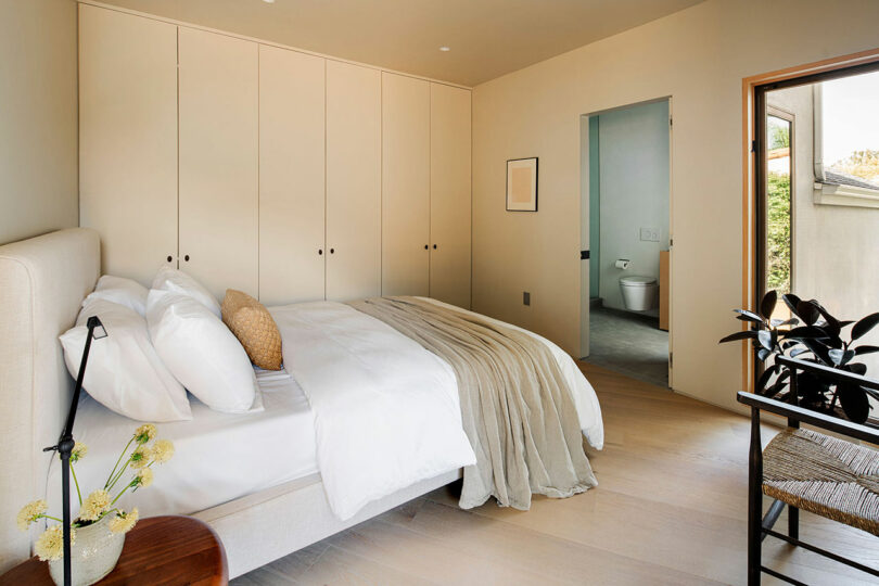 A modern bedroom featuring a bed with white linens, beige walls with built-in closets, an ensuite bathroom, a small wooden side table with flowers, and a black chair by a window.