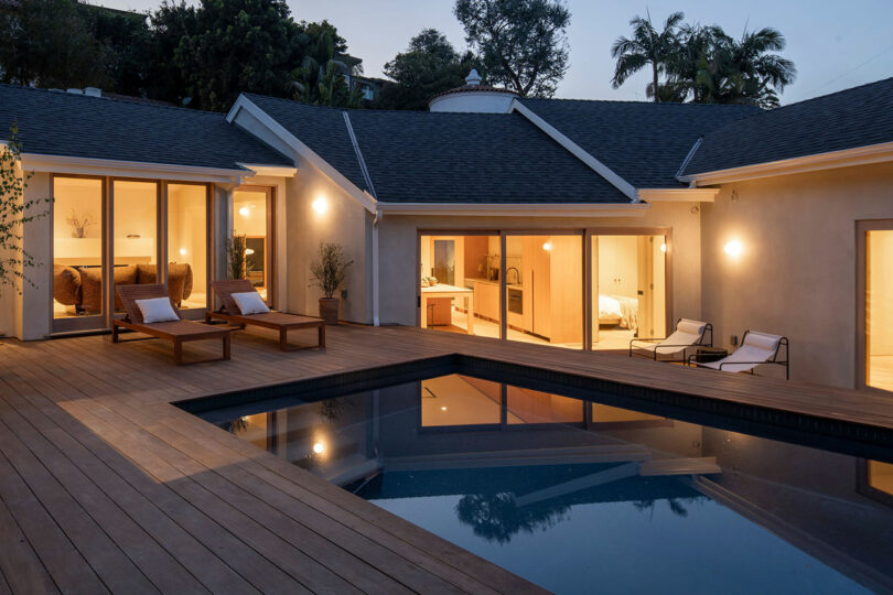 angled corner of a courtyard of a modern home's exterior at twilight with a wood deck surrounded pool and a lit house.