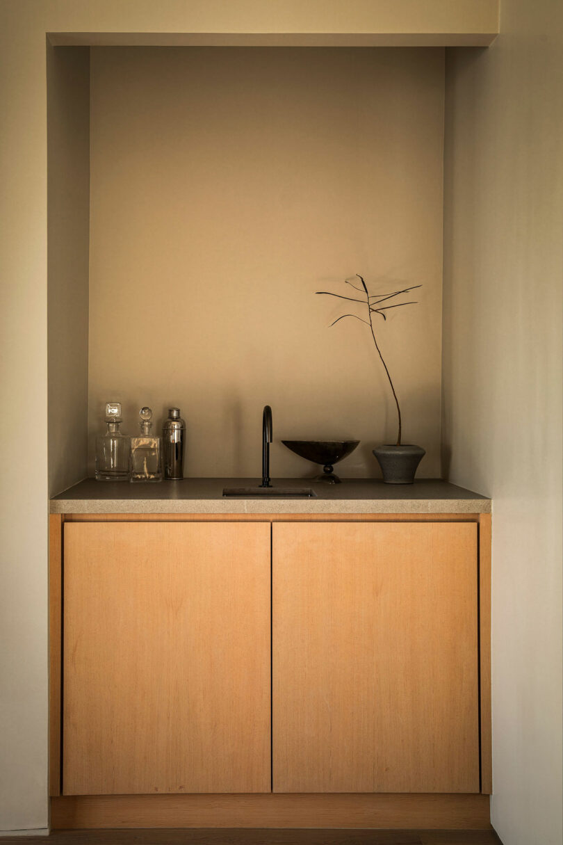 A minimalist nook with light wood cabinetry, a small sink with a black faucet, glass bottles, a cocktail shaker, and a decorative branch arrangement in a black vessel.