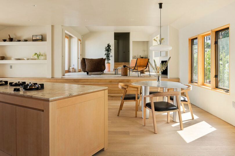 A modern, sunlit kitchen and dining area with light wood cabinetry, a round dining table, black chairs, and large windows. A two-step platform leads to a cozy seating area and a room with potted plants.