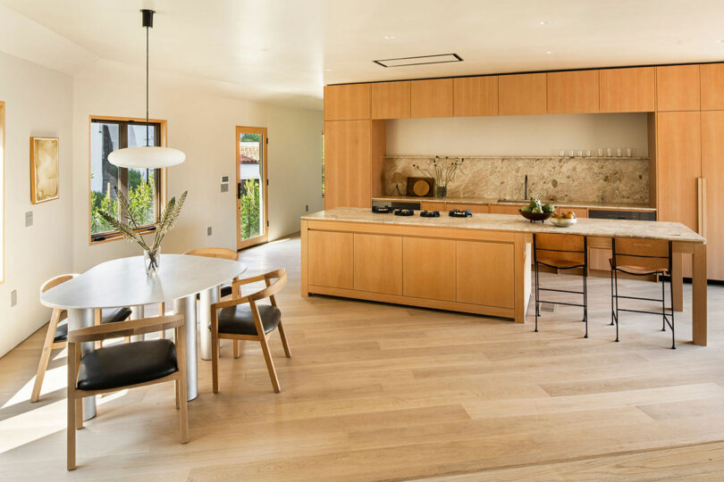 A modern kitchen with light wood cabinetry, a marble backsplash, and a central island with stools. A round dining table with four chairs is positioned to the left under a pendant light.