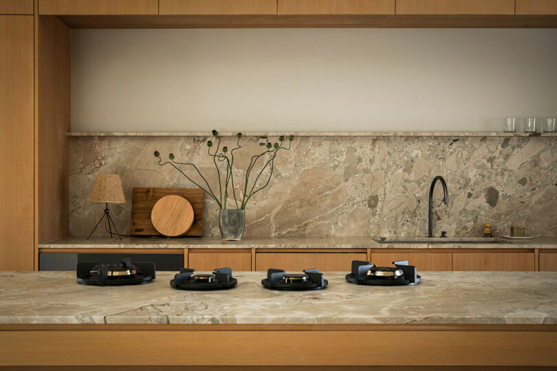 A modern kitchen with light wood cabinetry, a marble countertop and backsplash, a plant in a vase, kitchen utensils, and four identical black appliances lined up on the counter.
