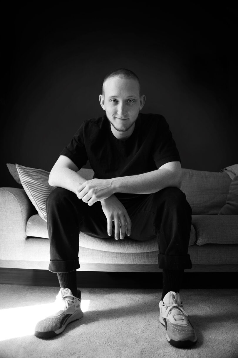 A person with a shaved head and casual attire sits on a couch, smiling and looking directly at the camera in a black and white photograph.