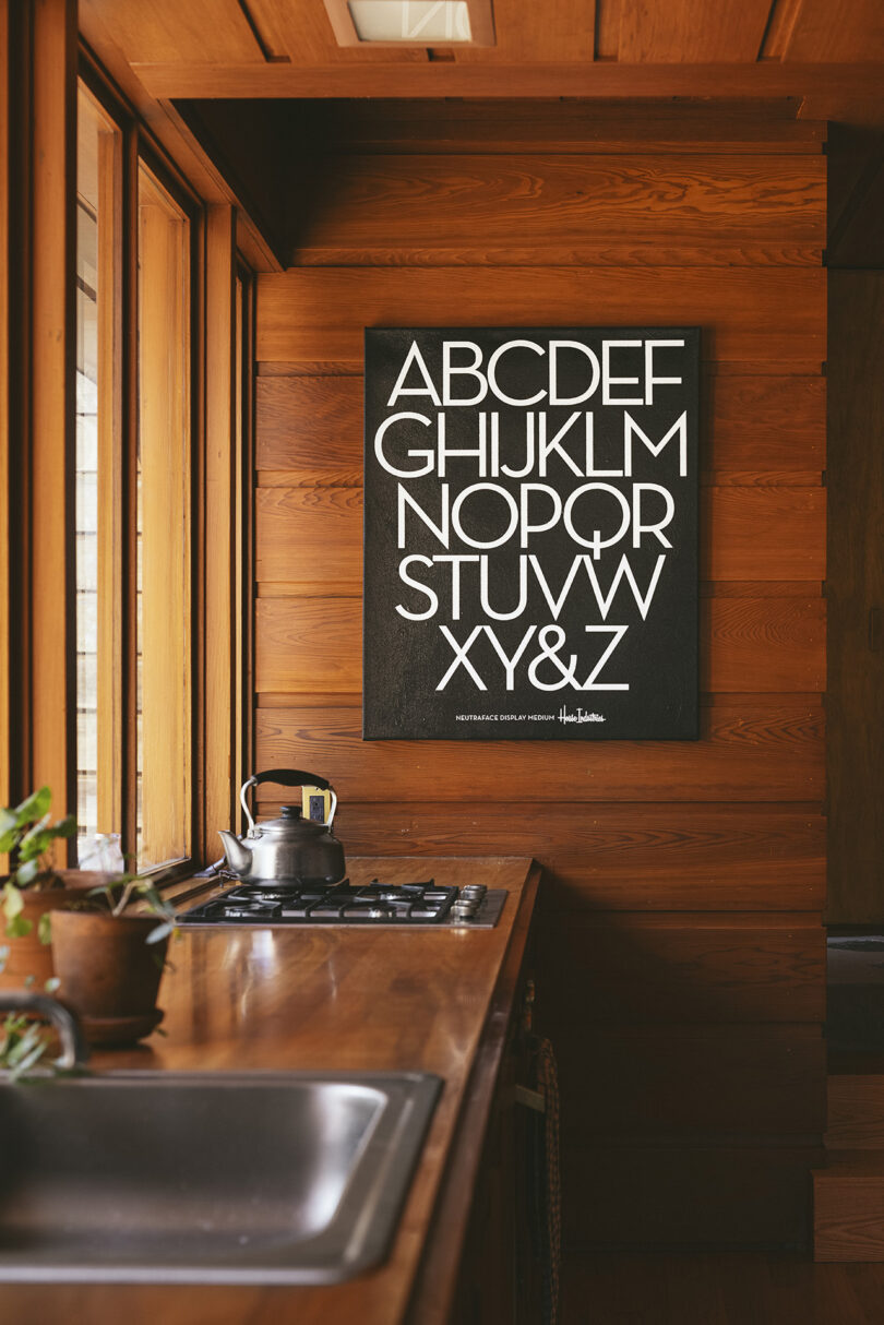 White on black serigraph of the alphabet in a modern font hanging on a wood paneled wall.