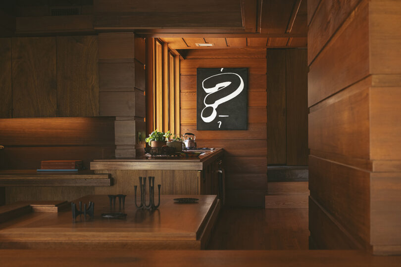 Wood-paneled room with sleek furniture, small plants, utensils hanging on the wall, and a minimalist art piece depicting a question mark.