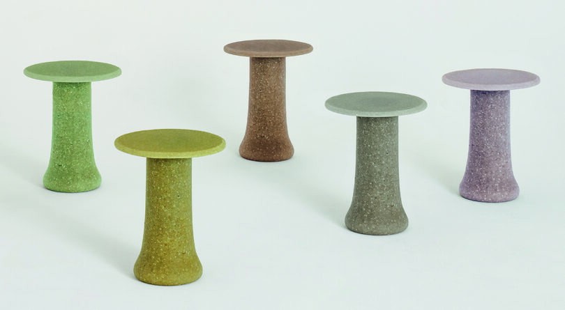 Five colorful, cylindrical side tables with flared bases are arranged in a scattered formation against a plain backdrop. The tables are green, yellow, brown, gray, and purple.