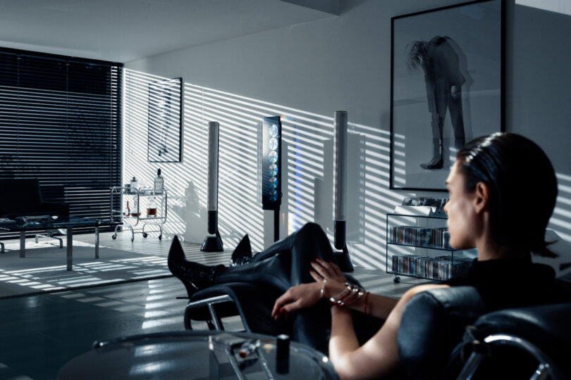 Woman with slicked back hair wearing black tights, tank top, and stiletto heel boots seated in lounge chair near Beosystem 9000c audio system, with light and shadows cast across a nearby wall.