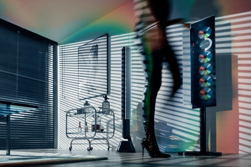 Woman wearing black tights, tank top, and stiletto heel boots standing near Beosystem 9000c audio system, with light and shadows cast across a nearby wall.