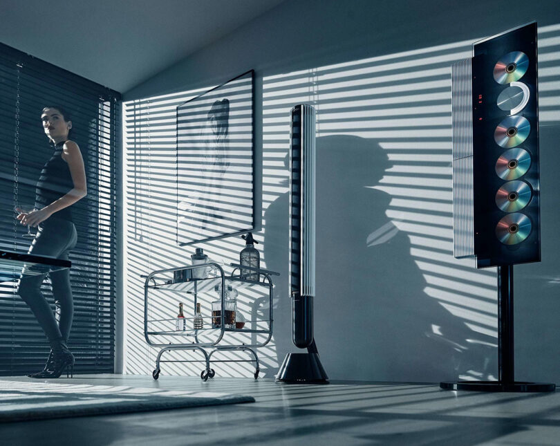 Woman with slicked back hair wearing black tights and tank top standing near partially opened blinds with light shining into the room and the Beosystem 9000c audio system, with shadows cast across the wall.