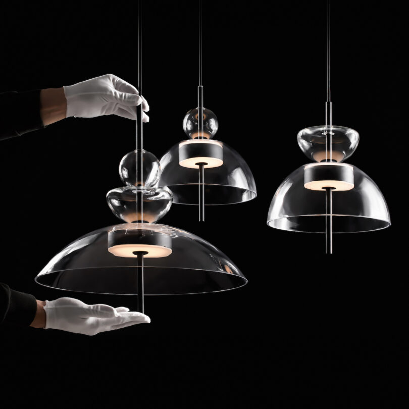 Gloved hands adjusting modern pendant lights with transparent glass covers and visible light bulbs against a dark background
