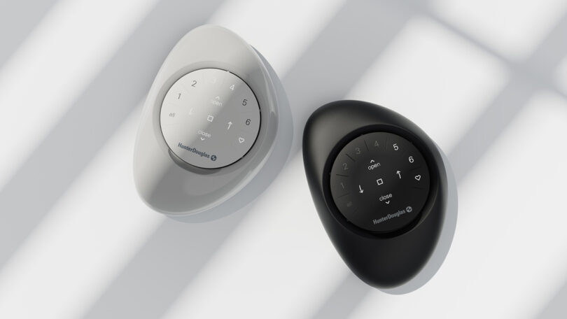 Two stone-shaped PowerView Pebble Remotes, one white and one black, are placed on a white surface with shadows from window panes.