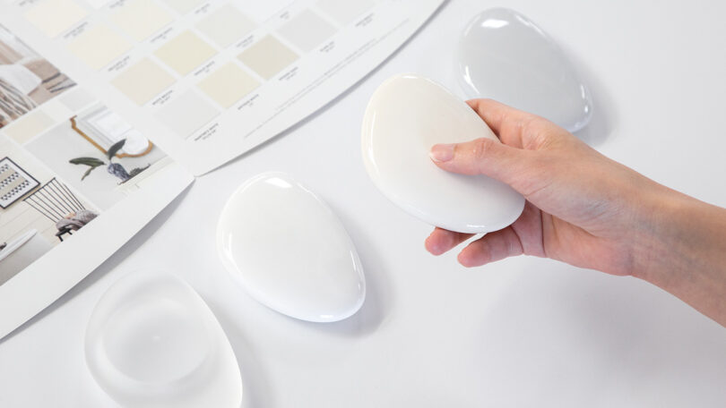 A hand holds an oval-shaped color sample, reminiscent of a stone. The PowerView Pebble Remote is surrounded by other similar design model samples and a color swatch booklet on a white surface.