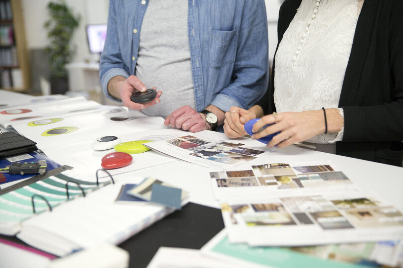 Two individuals stand at a desk reviewing magazines and color samples, with one wearing a denim shirt and the other a white laced top. Various office materials, sample swatches, and a PowerView Pebble Remote are spread out on the desk.