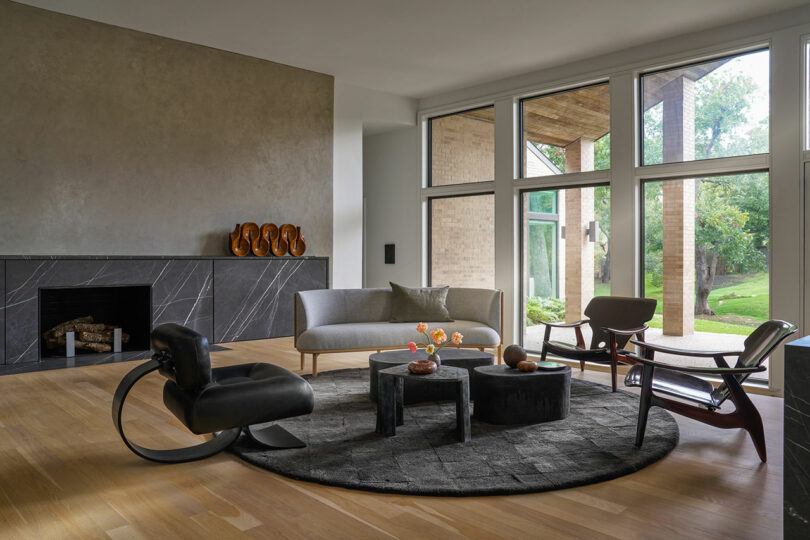 A modern living room with a fireplace, a gray sofa, unique armchairs, and a round rug. Large windows offer a view of the outdoor greenery. Inspired by Brazilian Modernism, this space features minimalistic decor and hardwood floors.