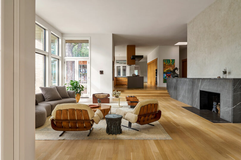 Modern living room featuring large windows, wooden floors, a gray fireplace, a sofa with throw pillows, two wooden lounge chairs with white cushions, and a central round coffee table that exudes Brazilian Modernism.