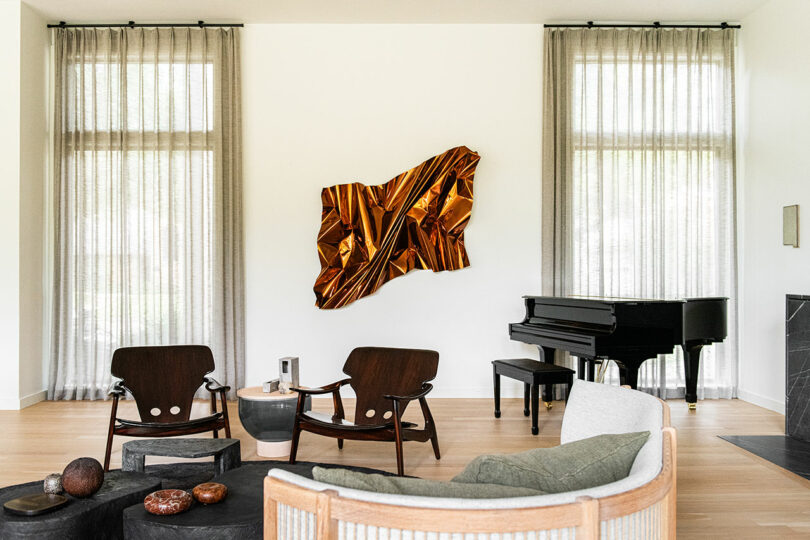 A modern living room inspired by Brazilian Modernism featuring gold abstract wall art, two wooden chairs, a wooden coffee table, a round green chair, a black grand piano, and large windows with sheer curtains.