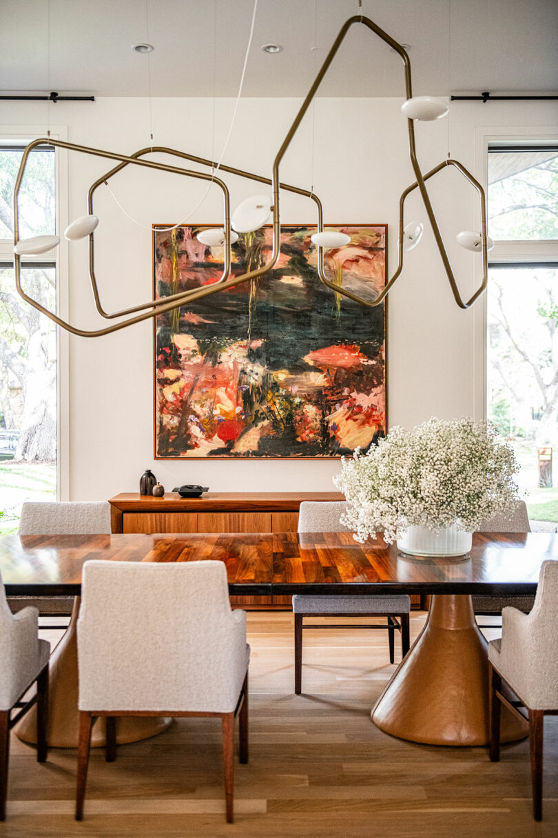 A modern dining room features a wooden table with white chairs, a large abstract painting on the wall, geometric light fixtures inspired by Brazilian Modernism, and a vase of white flowers.