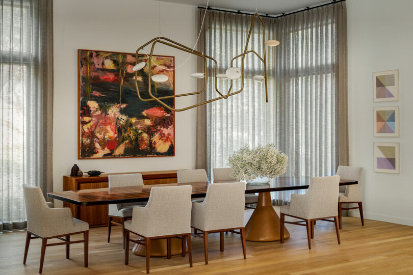 A modern dining room featuring a rectangular table, eight upholstered chairs, a contemporary chandelier, and floral centerpiece. The space is adorned with abstract wall art and large windows with sheer curtains, allowing light to dance on the wooden flooring, embodying elements of Brazilian Modernism.